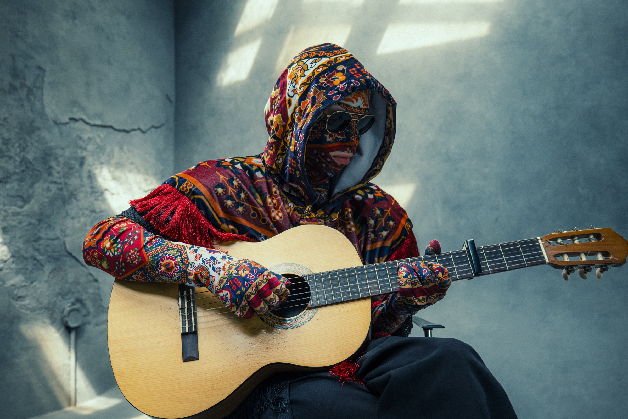 Carpetman — masked in traditional carpet patterns — playing an acoustic guitar in sunlight